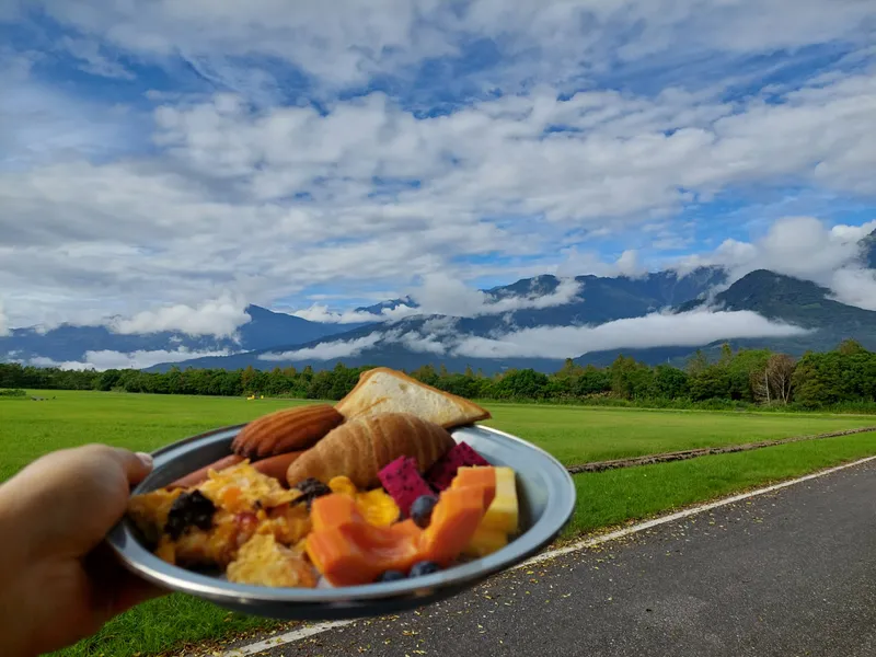 A hand holding a plate of bread and fruit in front of green fields and cloud-covered mountains