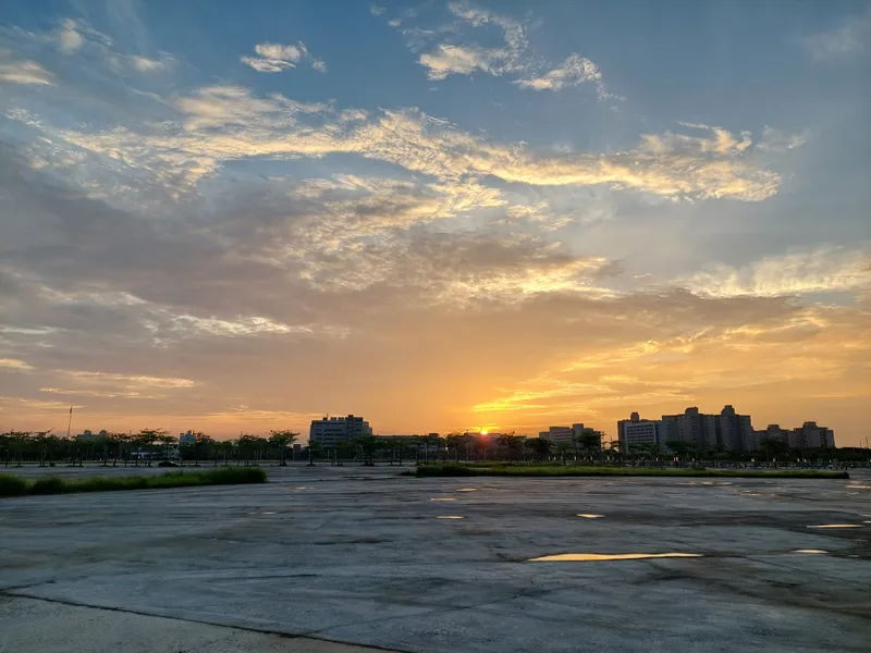 The sun setting between distant buildings under orange clouds, with puddles on the ground