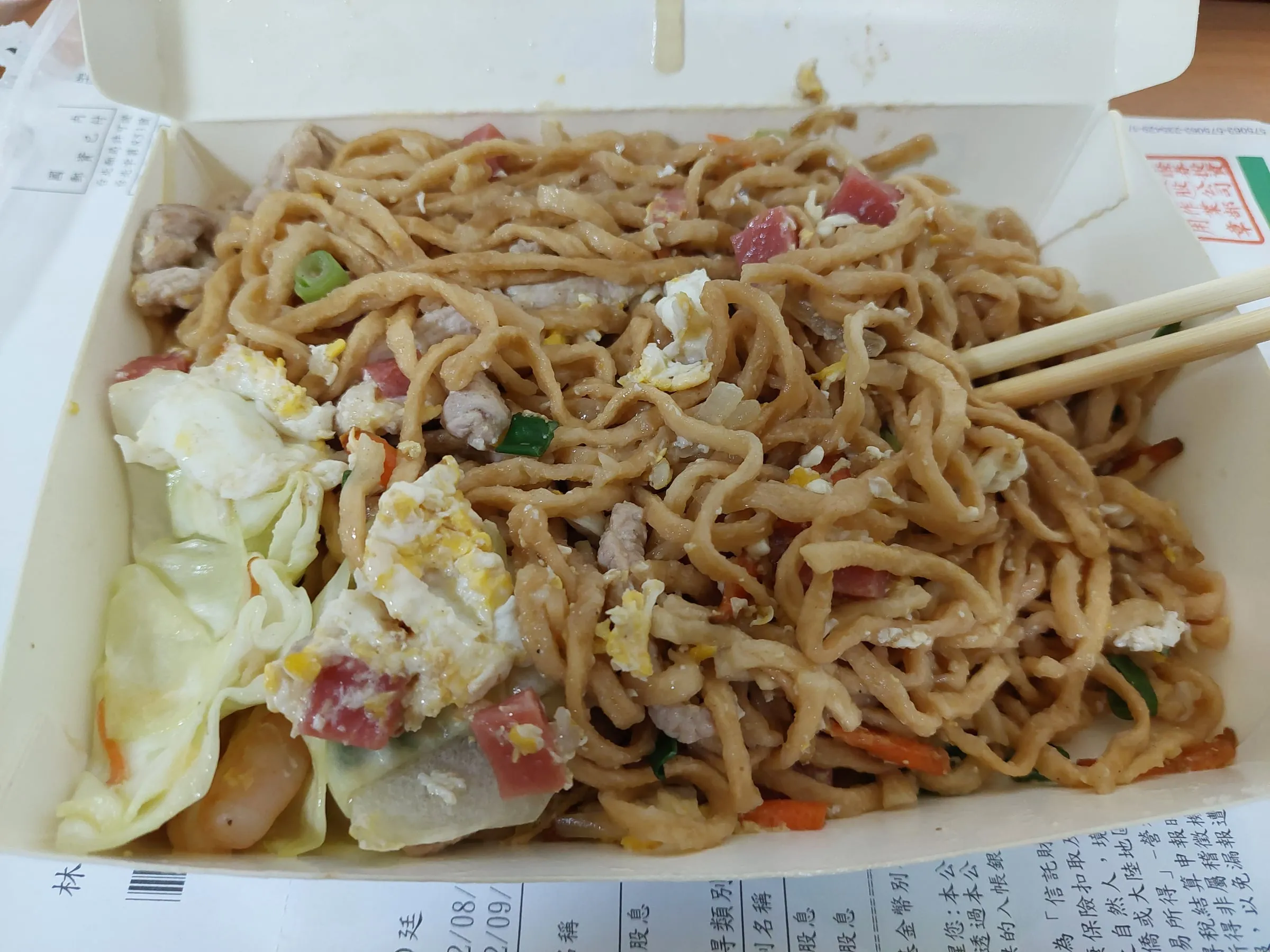 A takeout box full of fried noodles with cabbage, egg and diced meat