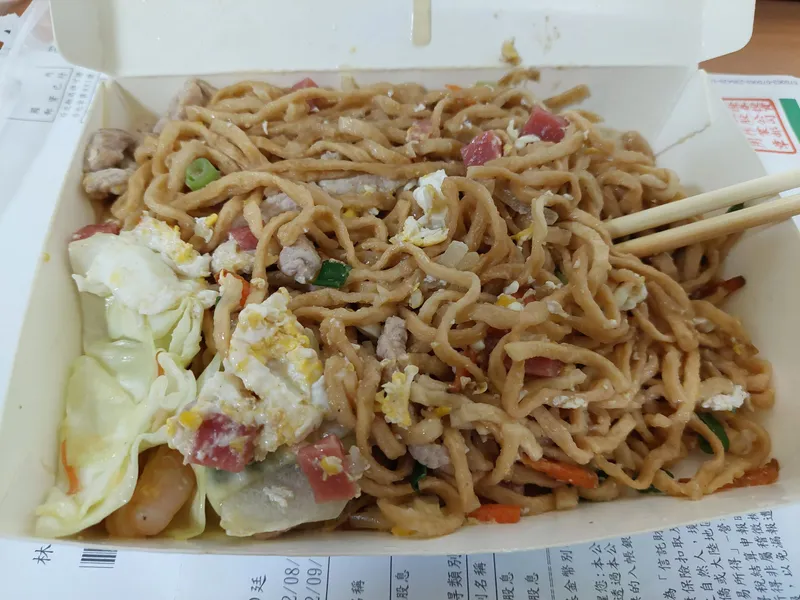 A takeout box full of fried noodles with cabbage, egg and diced meat