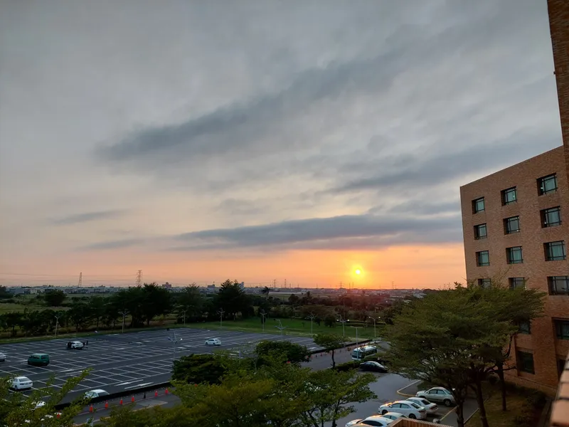 The sun setting over a distant plain, with a parking lot and trees in the foreground