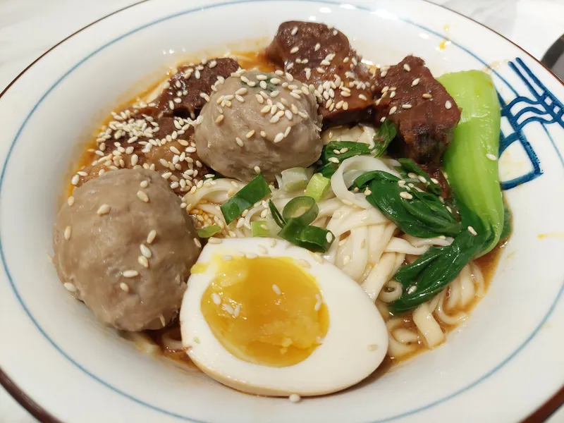 A bowl of beef noodle soup with meatballs, a soft-boiled egg and bok choy, sprinkled with sesame