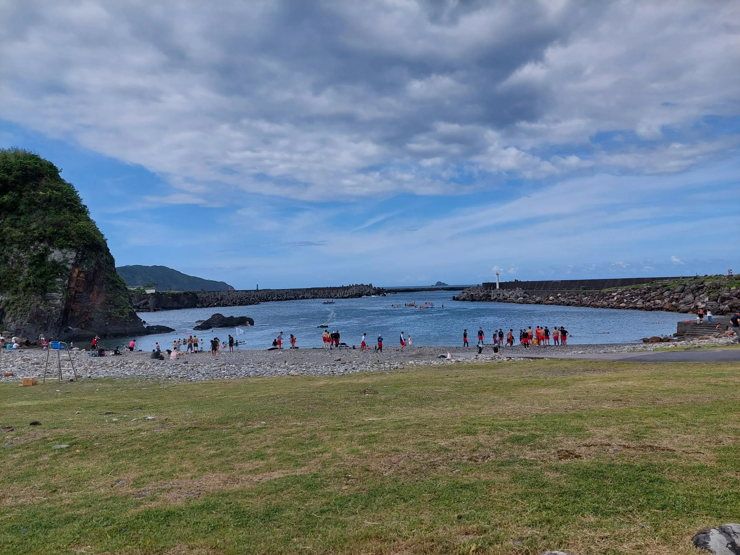 A bay beyond a grassy field, with many people along the shore and a rocky headland on the left