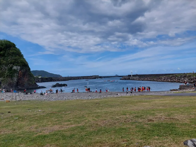 A bay beyond a grassy field, with many people along the shore and a rocky headland on the left