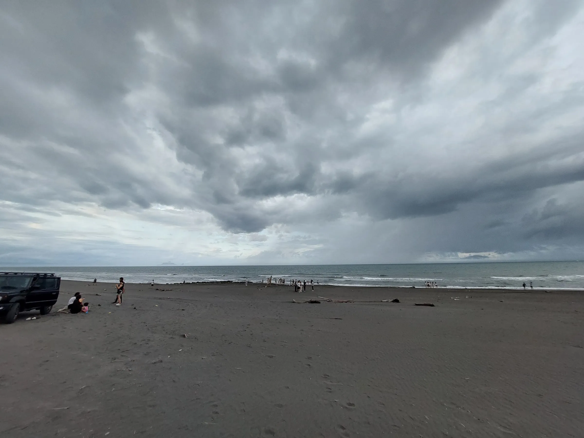 Heavy dark clouds over a grey sand beach, with a few people in the distance