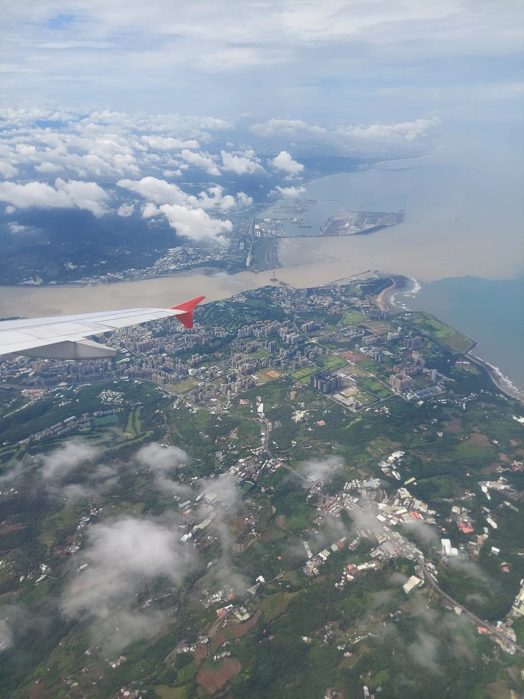 View from an airplane window showing the wing, clouds and a coastline below
