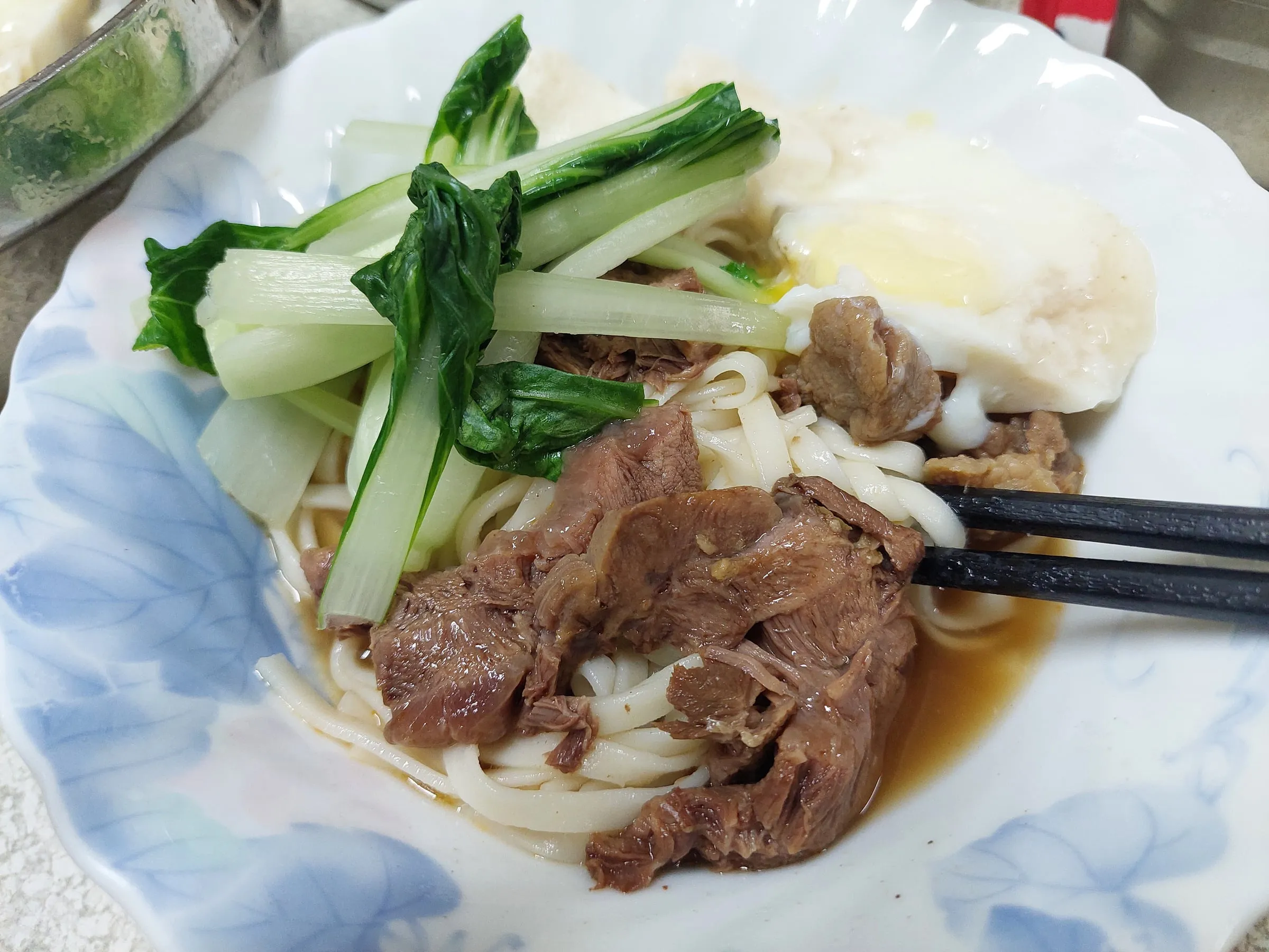 Noodles in a white porcelain dish, topped with sliced beef and bok choy