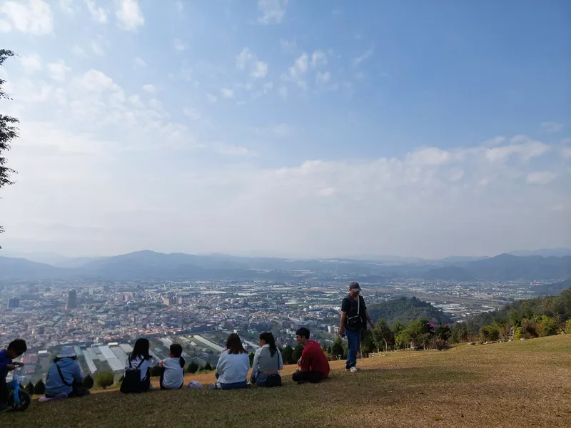 A few people sitting on a grassy hilltop, looking out over a city and distant mountains