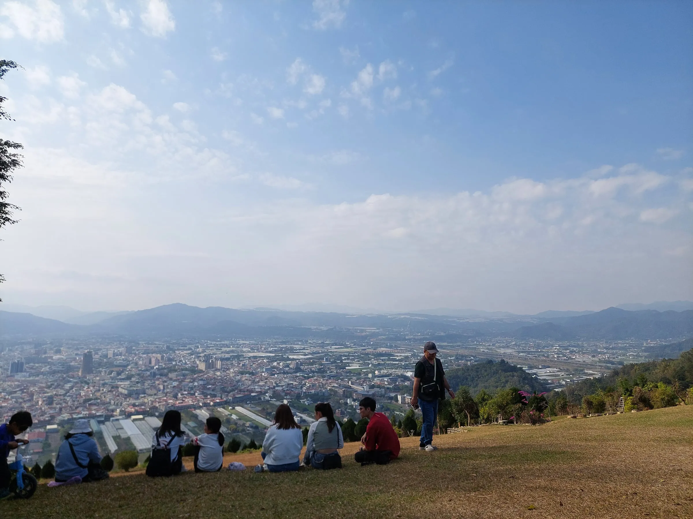A few people sitting on a grassy hilltop, looking out over a city and distant mountains