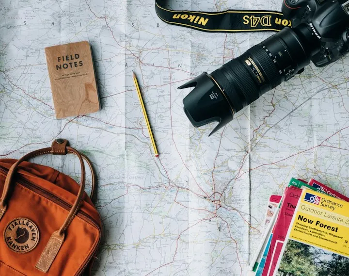 Overhead arrangement of a Nikon DSLR, orange Fjallraven backpack, Field Notes notebook, and maps on a paper topographic map