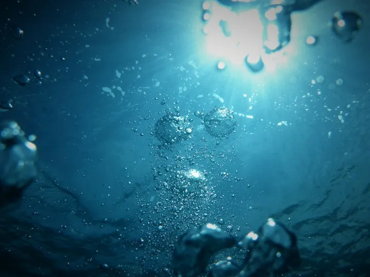Underwater view looking up at the sun with air bubbles rising through deep blue water