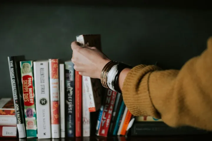 Hand in a mustard-yellow sweater reaching for a book on a dark shelf among crime and fiction titles
