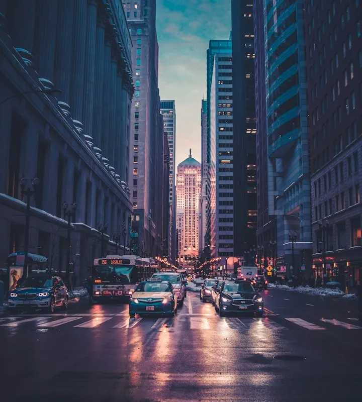 Vertical view down a wet city street at dusk with skyscrapers, a bus, and headlights reflecting on the pavement