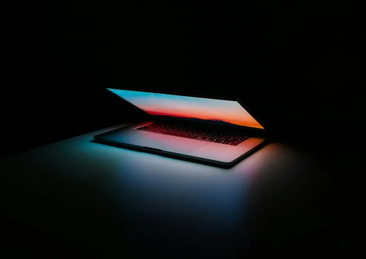 Partially open laptop in a dark room, its screen showing a sunset that lights the keyboard