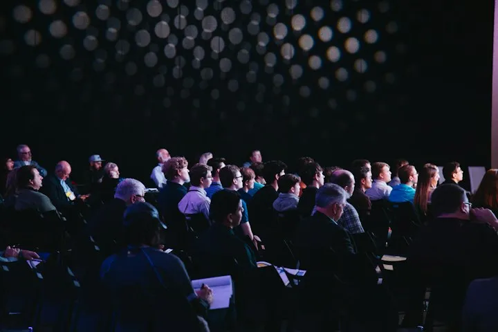 Seated audience in a darkened conference hall looking toward a presentation