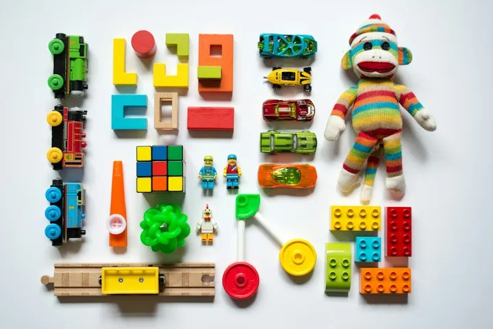 Overhead flat-lay of colorful children's toys including trains, cars, blocks, a Rubik's cube, and a sock monkey