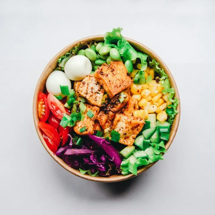 Top-down colorful Buddha bowl with seared tofu, corn, cucumber, red cabbage, tomatoes, and quail eggs