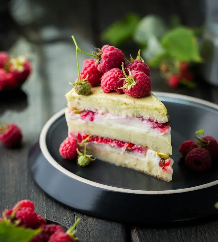 Slice of layered sponge cake topped with fresh raspberries on a dark plate